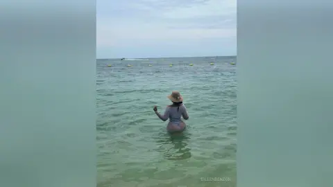 Woman in a grey swimsuit and sun hat wading in the calm turquoise ocean water on a sunny day.