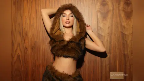 Blonde woman in a brown faux fur hooded crop top and skirt posing against a warm wood paneled wall.