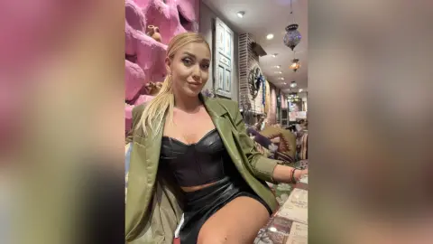 AmaFox — Blonde woman in a black leather outfit and olive green blazer sitting in a cozy, bohemian-style cafe.