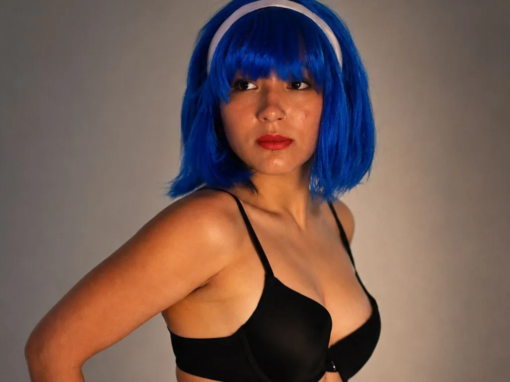 Cliofioreth poses in a black bra with bright blue hair against a plain grey background.
