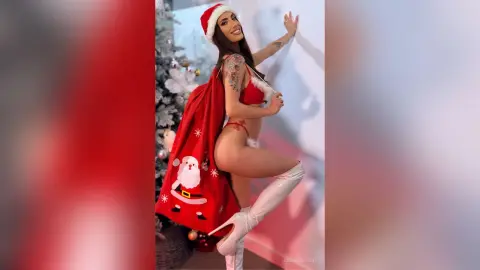 Tattooed woman in red Christmas lingerie and Santa hat posing playfully by a festive holiday tree.