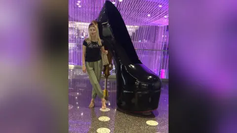 Woman in black t-shirt and olive trousers standing next to a giant black high heel shoe in a modern mall.