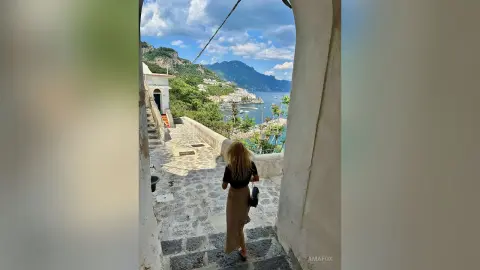 AmaFox — Blonde woman in a black top and tan skirt walking down stone stairs overlooking a scenic coastal Italian village.