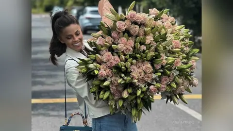 Smiling woman in a white shirt holding a massive bouquet of pink lilies on a city street.