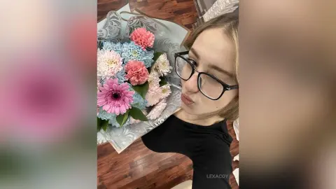 Young woman in a black top posing with a colorful bouquet of pink and blue flowers in a bright indoor setting.