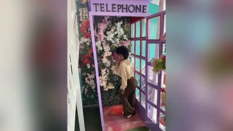 Young woman in a beige sweatshirt posing by a floral wall inside a vintage pink telephone booth.