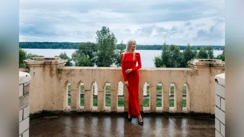 Elegant woman in a long red evening gown standing on a stone balcony overlooking a scenic lake at dusk.