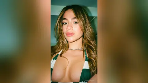 Young woman with long wavy hair wearing a green and white striped halter top in a soft indoor light setting.