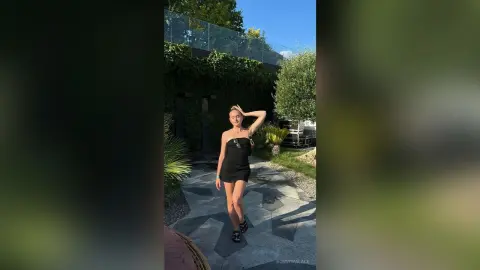 BonnyWalace poses in a short black dress outdoors on a stone path near green plants and trees.
