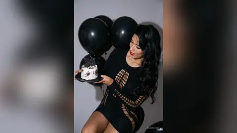 Woman in black cutout dress holding a birthday cake with black balloons in a celebratory mood.