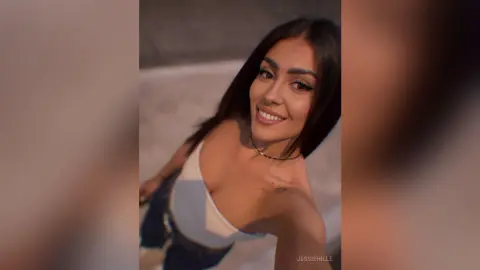Smiling young woman in a white tank top and jeans posing for a casual selfie in a bright indoor setting.