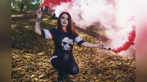 Red-haired woman in a black graphic t-shirt holding pink smoke bombs in an autumn forest setting.