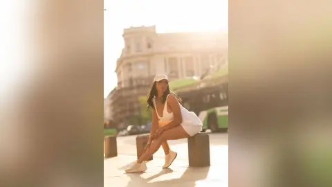 NicoleBella sits on a stone bollard wearing a white tank top and white shorts with a white cap.