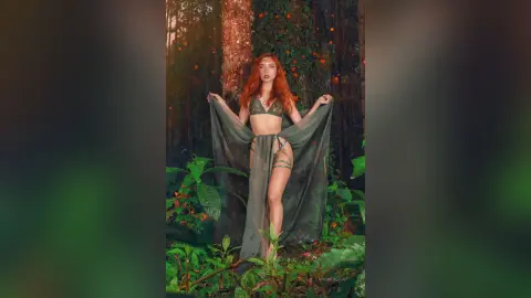 LaurentBelluci stands in a forest wearing a sheer green lace bra and matching thong, holding up a long, flowing translucent skirt.