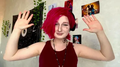 Woman with bright pink hair and black tank top smiling in a bedroom decorated with posters and vines.