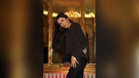 Elegant woman in a black blazer posing in a luxurious gold-framed mirror setting with warm lighting.