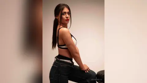 Young woman with long dark hair in a white sports bra and denim jeans posing confidently against a plain wall.