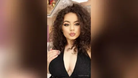 Glamorous woman with voluminous curly hair wearing a black lace plunging neckline dress in a soft indoor setting.