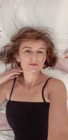 Young woman in a black tank top relaxing on white bedding with a calm, serene expression.