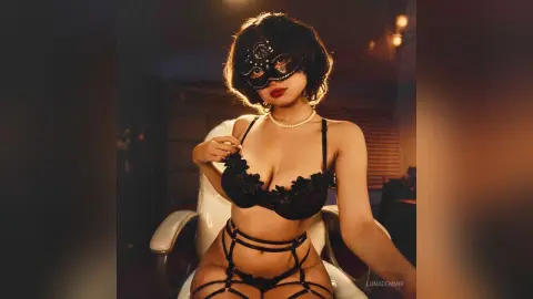 Glamorous woman in black lace lingerie and masquerade mask sitting in a moody, dimly lit room.