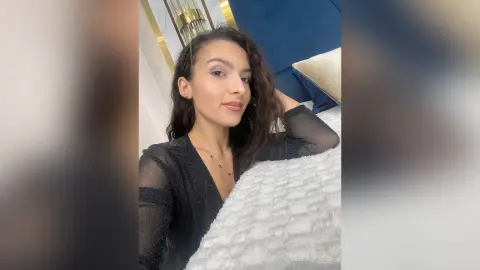 Elegant woman in a black sheer long-sleeve top relaxing on a white textured bed in a modern bedroom.