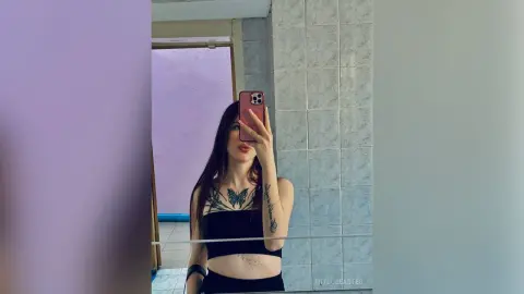 MitsueEastes poses for a mirror selfie wearing a black crop top, showing off her long dark hair and butterfly tattoo on her chest.