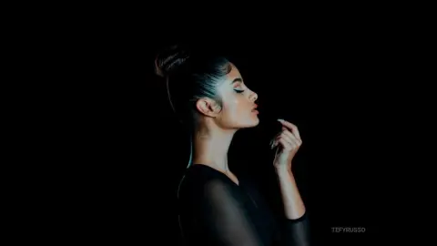 TefyRusso poses in profile against a black background, wearing a black sheer long-sleeve top with her hair pulled back in a tight bun.