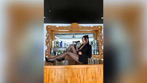 Elegant woman in a black blazer and patterned tights sitting on a wooden bar in a moody lounge setting.