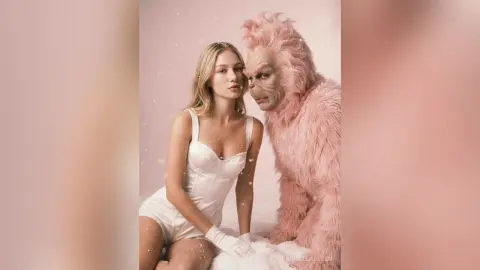 Blonde woman in a white satin bodysuit posing with a pink furry creature in a dreamy, soft pink studio setting.