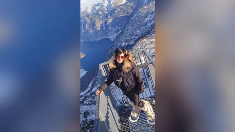 Woman in black winter jacket and sunglasses standing on a mountain balcony overlooking a snowy alpine lake.