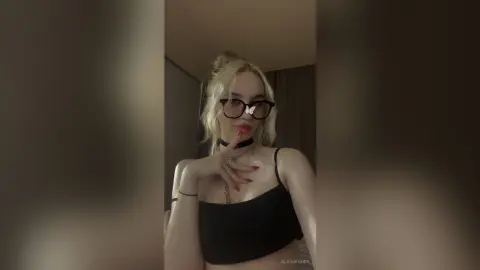 Blonde woman in a black crop top and glasses posing with a moody aesthetic in a dimly lit room.