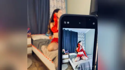 Young woman in a red outfit sitting on a bed in a cozy bedroom, captured via smartphone screen.