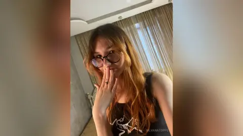 Young woman with long reddish hair and glasses wearing a black tank top in a bright, modern room.