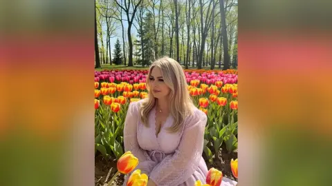 Blonde woman in a light pink lace dress smiling in a vibrant orange tulip field under sunny spring trees.