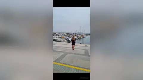 Woman in a black dress standing on a concrete pier by luxury yachts in a serene marina setting.