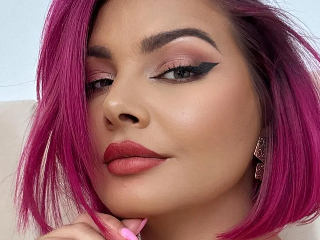 Woman with vibrant pink bob hair and dramatic winged eyeliner makeup looking confident in a bright setting.
