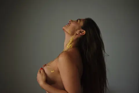 Smiling woman with gold glitter on her skin posing against a grey background in a dreamy, artistic mood.