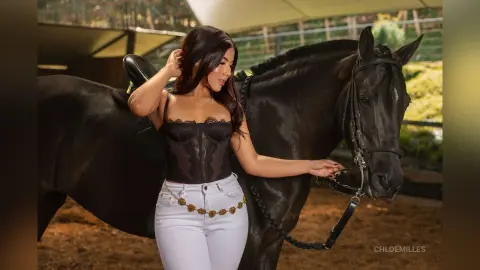 Elegant woman in a black lace corset and white jeans leaning against a dark horse in a rustic stable setting.