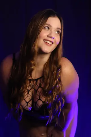 Smiling brunette woman in a black fishnet bodysuit posing against a dark blue studio background.