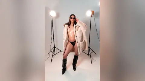 Fashionable woman in an oversized cream coat and black sunglasses posing in a bright photo studio.