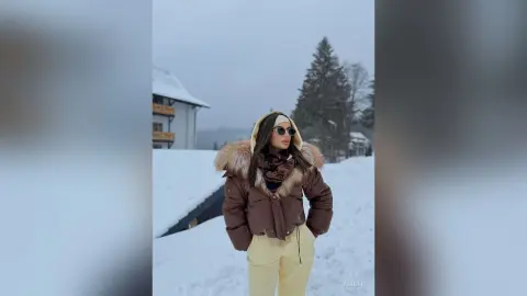 Woman in a brown puffer jacket and sunglasses standing in a snowy winter landscape.