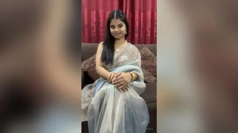 Sejuti wears a light grey saree and pearl necklace, sitting on a brown sofa against red curtains with her long black hair down.