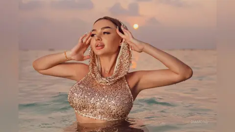 Glamorous woman in a gold sequin halter top posing in the ocean during a dreamy sunset.