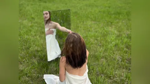Young woman in a white dress looking into a mirror in a lush green meadow during a dreamy outdoor photoshoot.