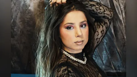 Woman with long wavy hair wearing a black lace top and silver chain necklace in a moody studio setting.