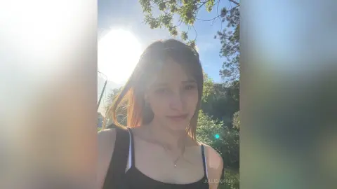 Young woman in a black tank top smiling in sunny outdoor garden setting with natural sunlight.
