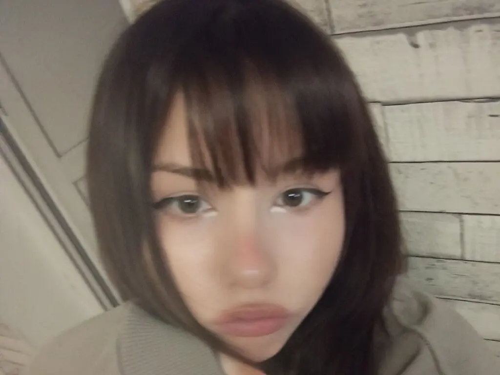 Young woman with brown hair and winged eyeliner wearing a neutral sweater in a cozy indoor setting.