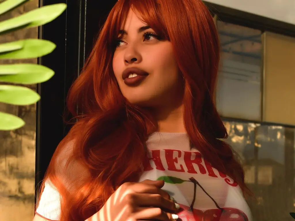 Redheaded woman in a white cherry print top posing outdoors in warm sunset lighting.
