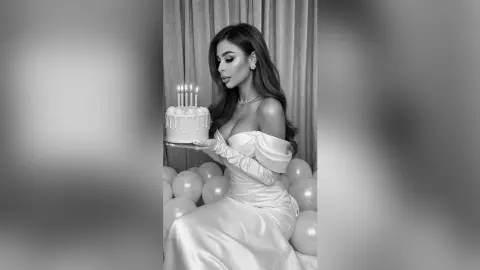 Elegant woman in a white satin gown holding a lit birthday cake in a dreamy, monochrome setting.