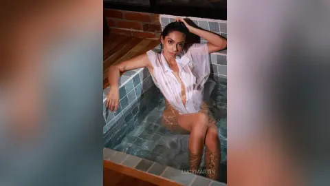 LucyMartin sits in a tiled sink or small tub, wearing a white button-down shirt that is partially open or wet.